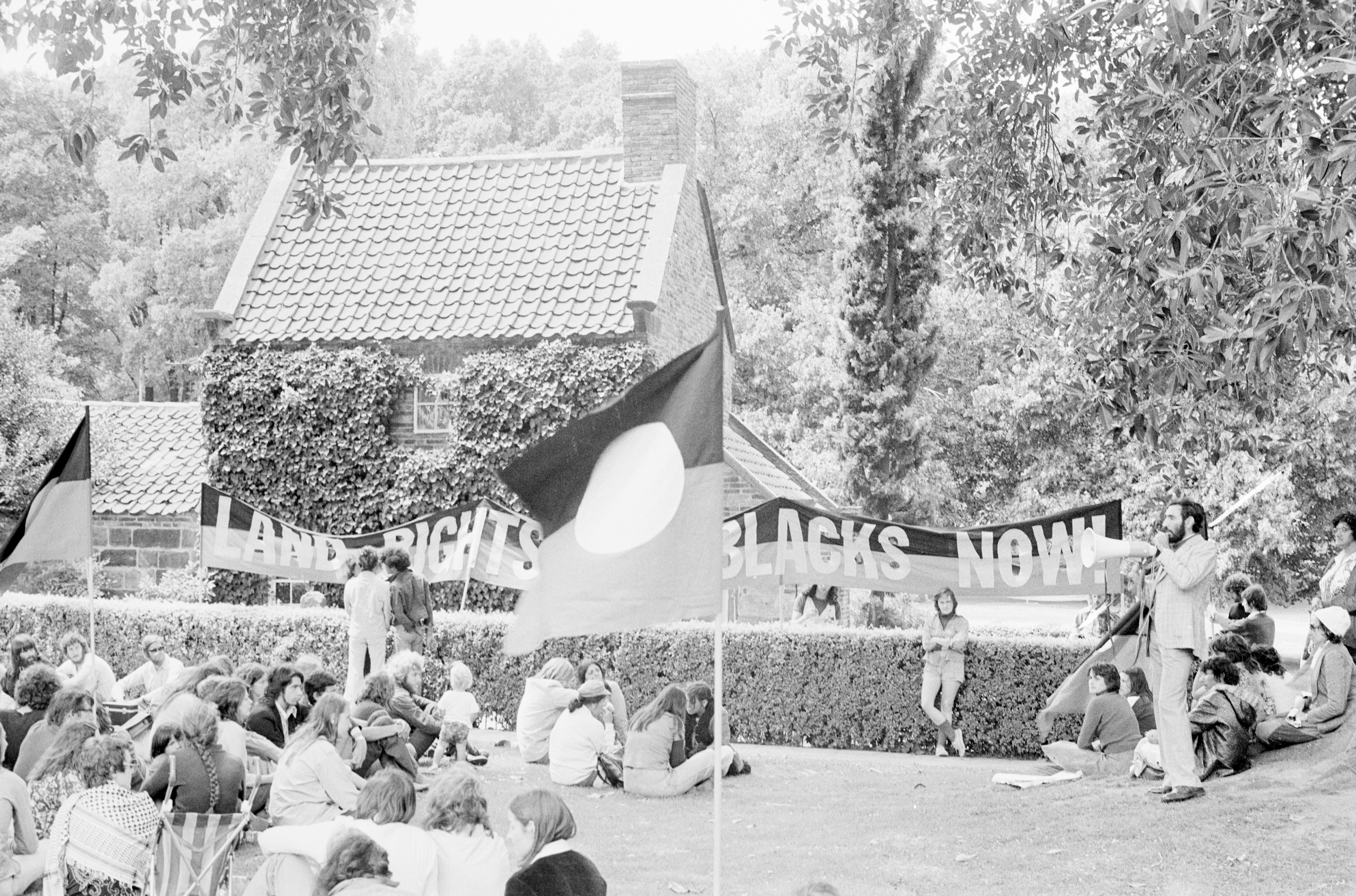 Black and white photograph of group of people gathered outside Cooks Cottage, in the Fitzroy Gardens, protesissues of land rights and uranium mining.Australian Aboriginal flags and banners surround Cook's cottage.