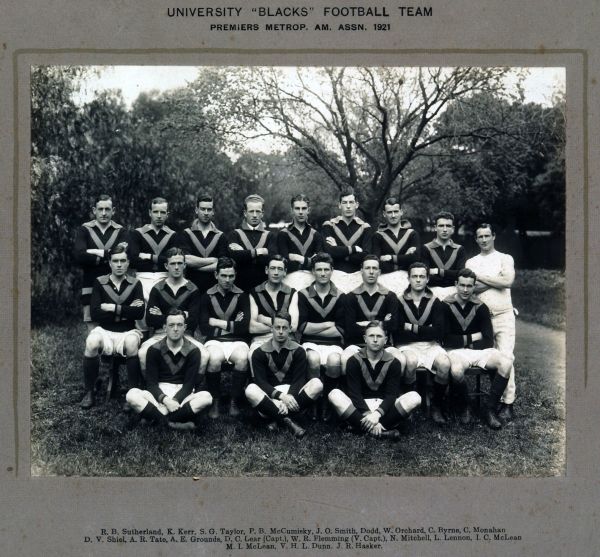Black and white photograph of men in football uniform of shorts, jumper with 'v' design, socks and boots, posing in three rows outdoors on grass area surrounded by trees, photogrpah mounted on grey card, title 'University 'Blacks' Football Team Premiers Metrop, AM. ASSN. 192 printed on card above photograph, names of players printed on card below photograph.