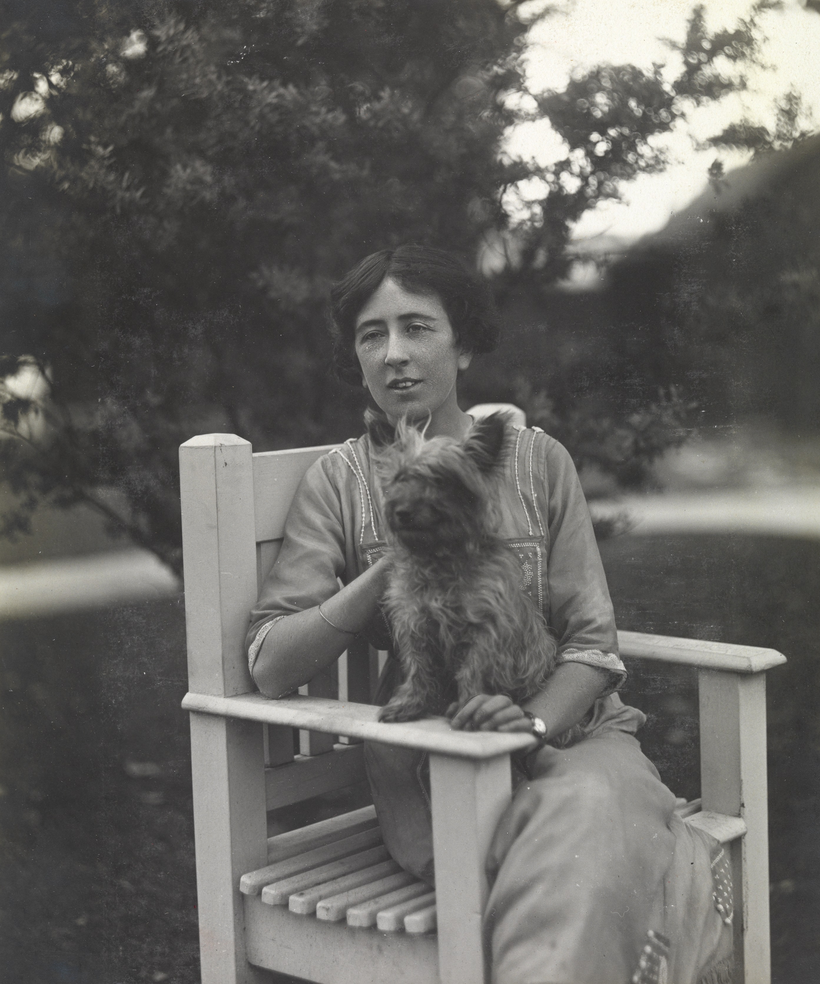 Woman and terrier dog sitting on garden chair both with gaze averted t their right  . terrier dog on her lap with front paws on arm of chair 