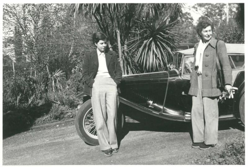 Black and white photograph of two women standing against vintage car.