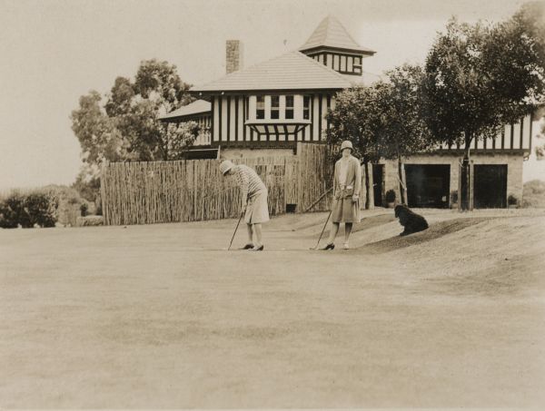 two women playing golf on grass field. Two story house with turret behind them. Dog sitting on grass slope under tree to the right. 