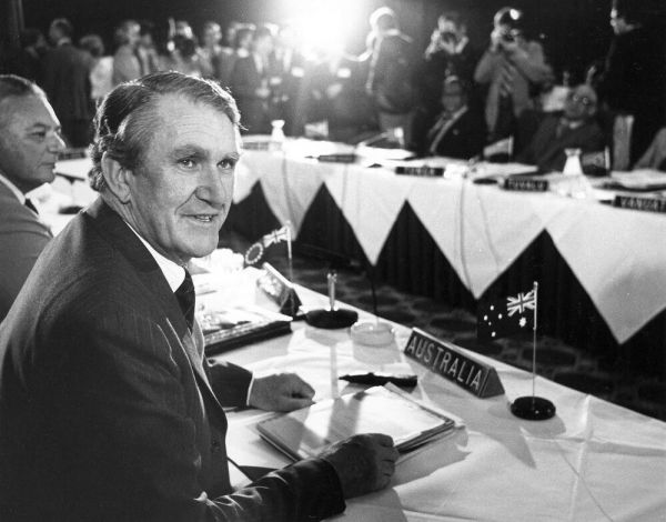 Black and white photograph of a man seated at a table with 'Australia' table marker, surrounded by people in formal attire.