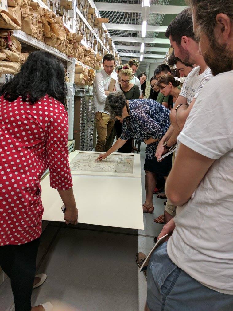 Group of people gathered around an open shallow drawer of paper records in large repository storage space
