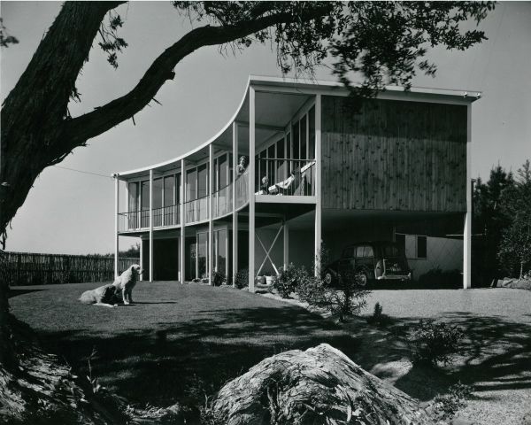 Black and white photograph of a two level modernist suburban home, ‘Gibson House’, framed by a tree, with two dogs sitting on on lawn in front of house, woman child on bacony of second level. 