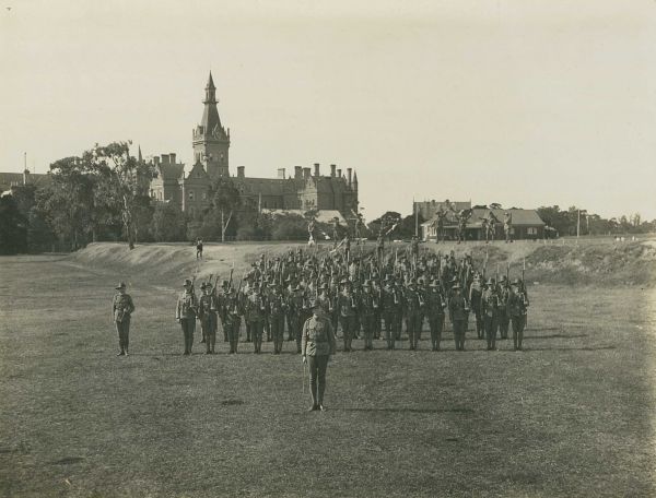 Black and white photogrpah of Rifles Club in uniform and in formation on grass field, Ormond college in distance 