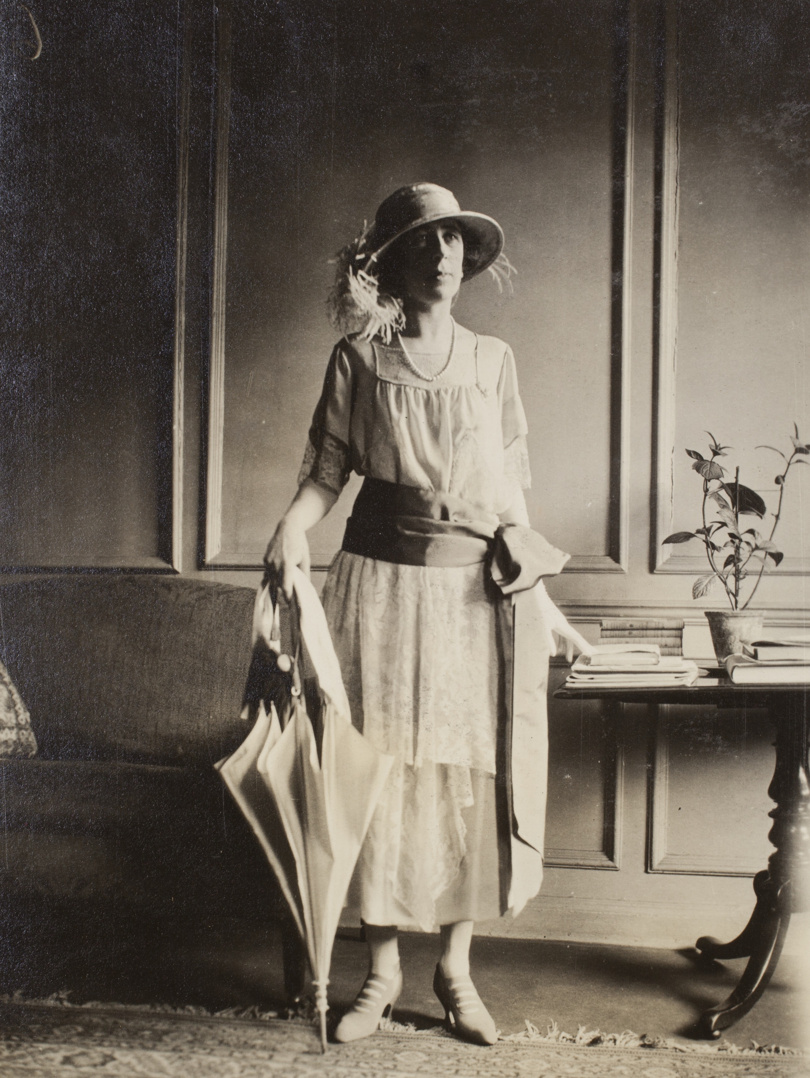 Woman standing in furnished room,dressed in summer frock with wideg ribbon tied into a bow side of waist, pearl necklace, hat with feather adornment, and holding half closed umbrella tip on ground 