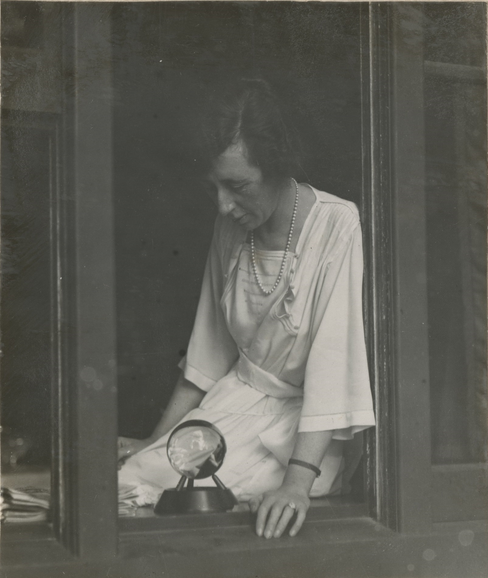 Woman dressed in white dress and pearl necklace  sitting on window ledge looking down at a clear glass ball on a stand