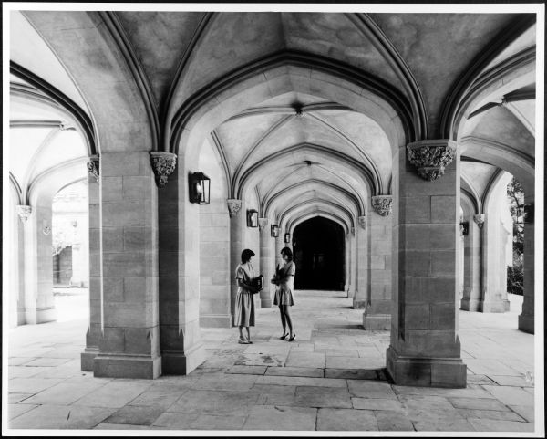 Black ansd white photograph of two women standing under building cloisters facing each other, both hugging folders, under building cloisters