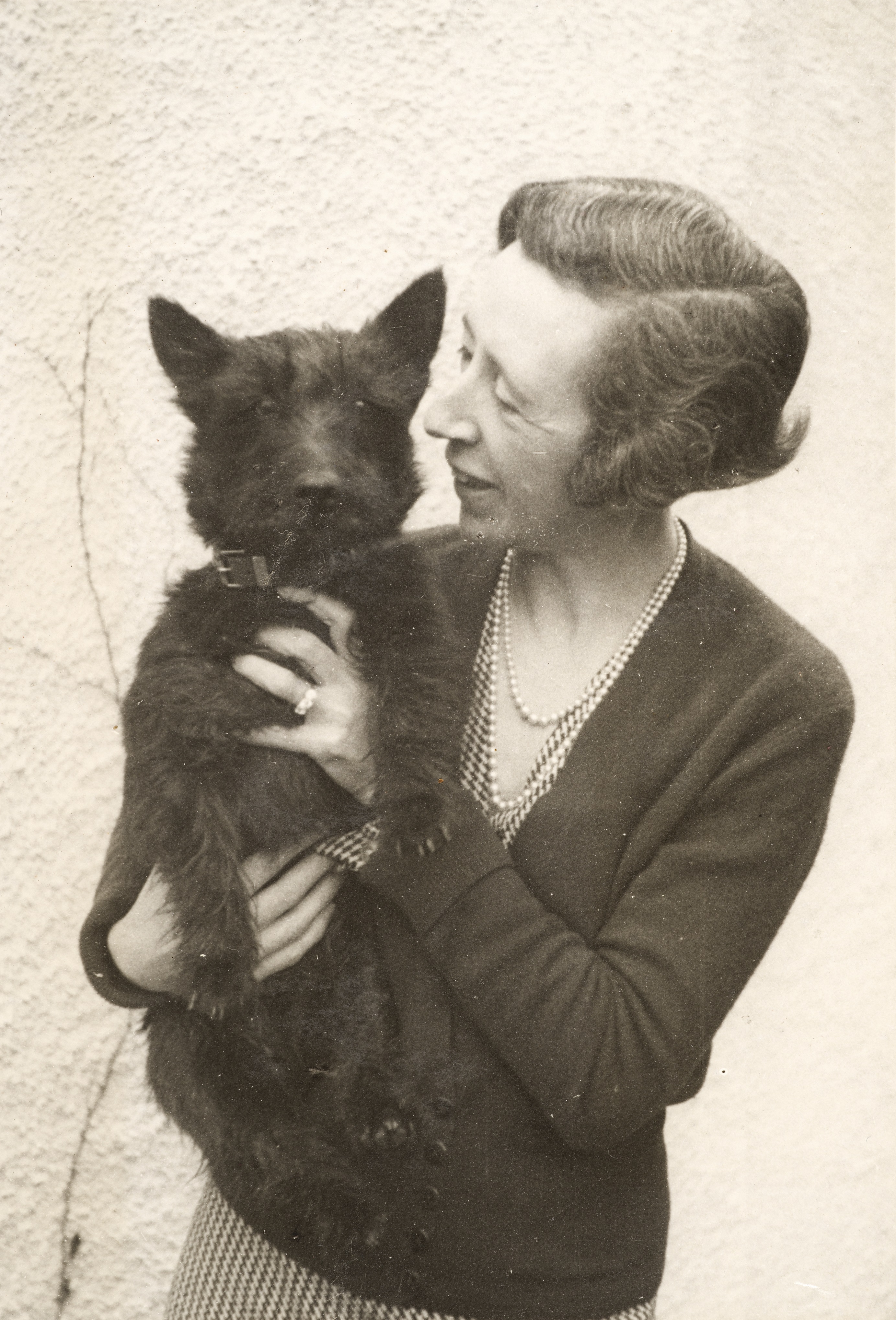 Woman dressed in check dress, cardigan and pearl necklace holding and smiling at a black Scottish Terrior dog 