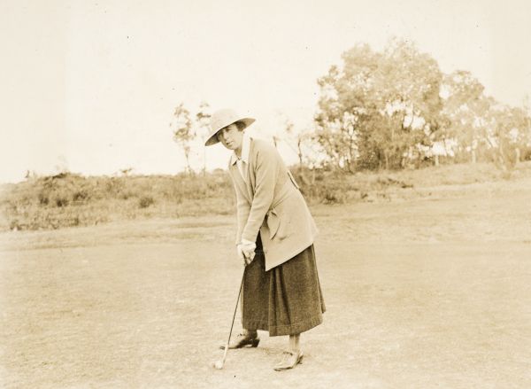 Woman dressed in long wool skirt, long cardigan and wide brim hat, standing in a golf putting stance, front on, holding putter