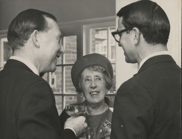 Two men in suits with backs to camera talking to older woman, all smiling, all holding drinking glasses 
