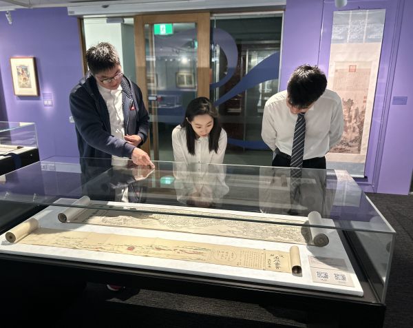 Three young people gathered around an ancient scroll in a long glass display cabinet. 