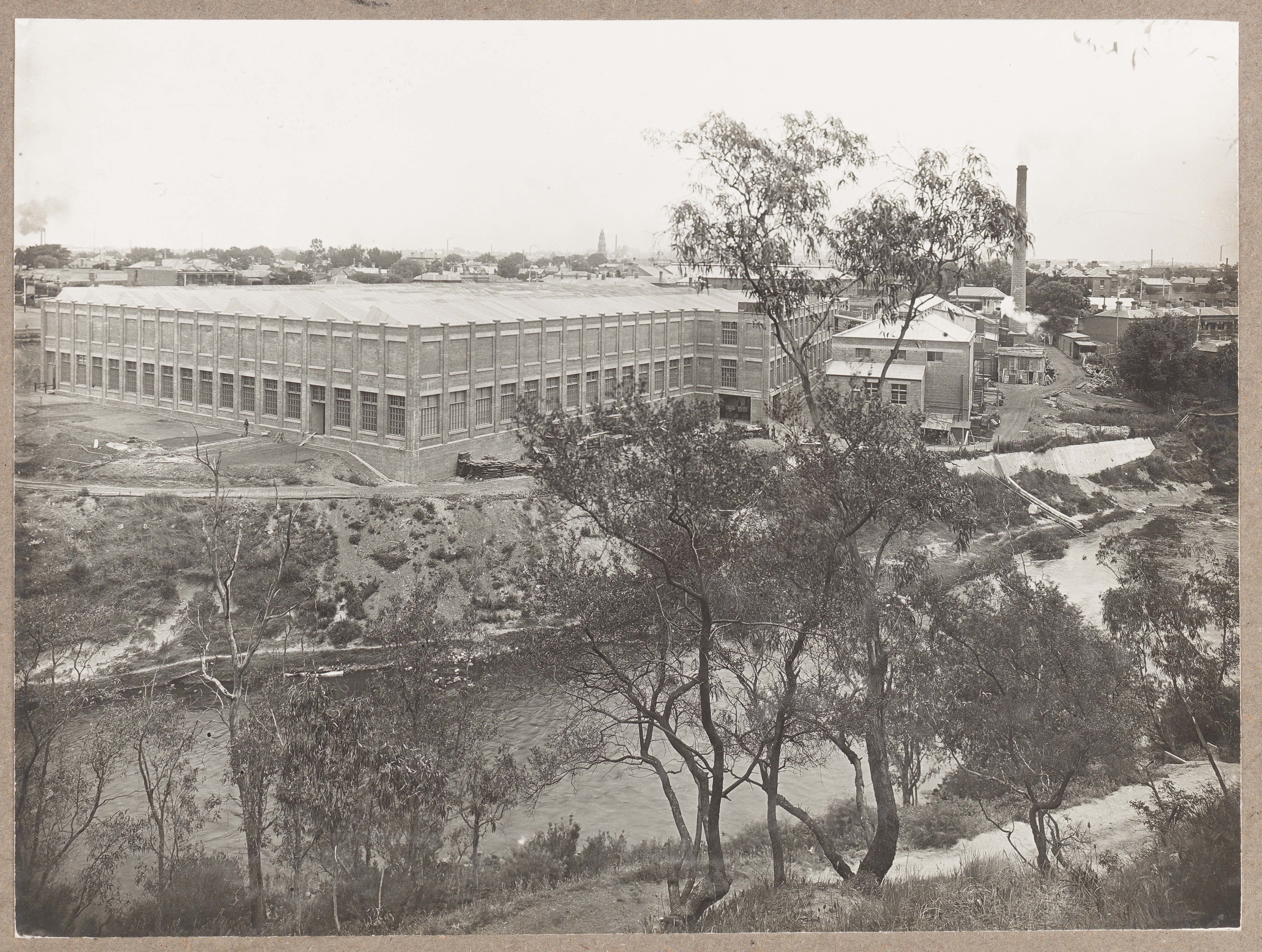 Black and white photograph of a large factory, with river in foreground surrounded by trees