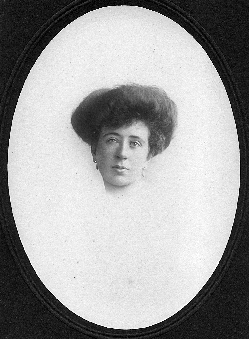 Photographic portrait of a white skinned woman, hair tied up in the Edwardian full pompadour style, high, rounded and curved away from the , wearing pearl teardrop earrings