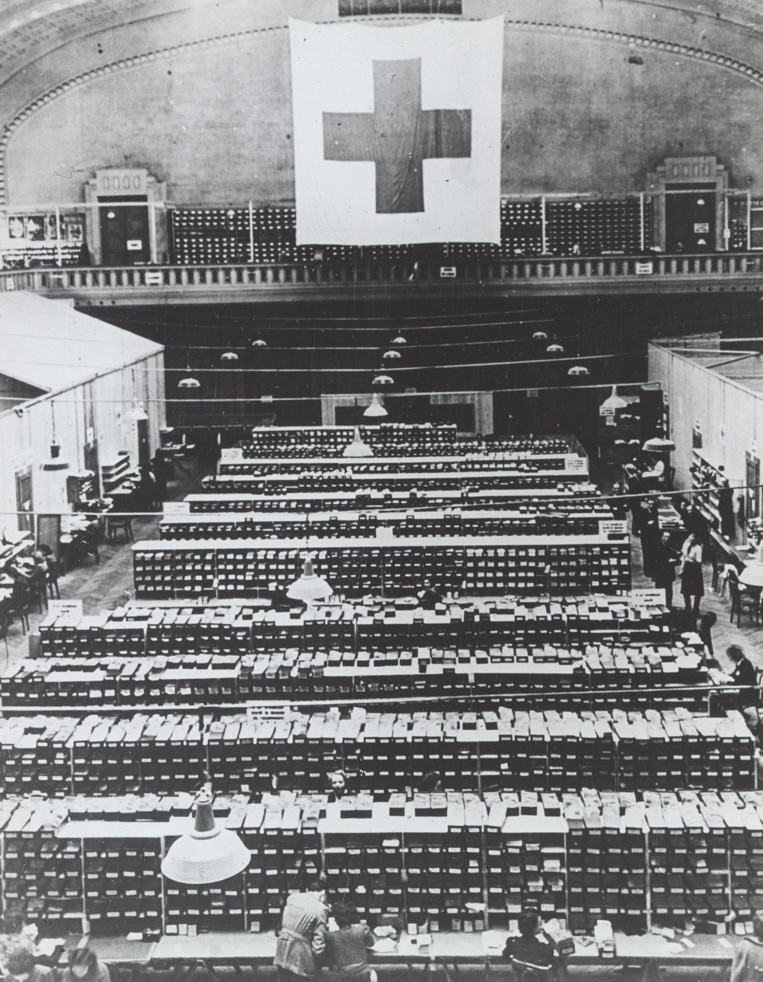 Aerial view of large hall filled with filing draws holding POW cards, large Red Cross Banner hanging above mezzanine at rear