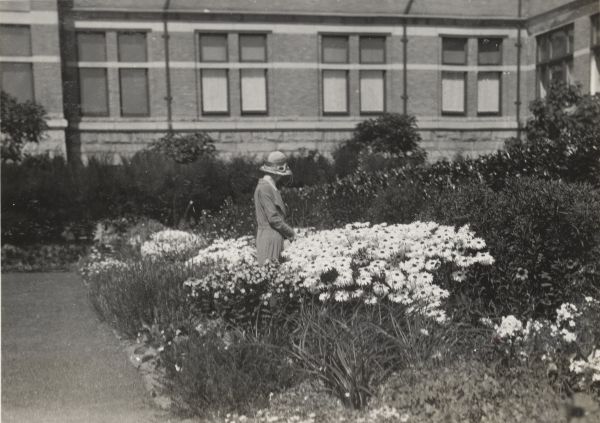 Woman wearing long coat and hat, face turned away,  standing in a garden of tall daisies in front of a stone and brick hotel